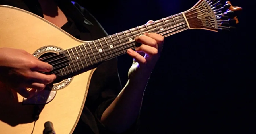 Mãos a tocar uma guitarra portuguesa de doze cordas em forma de pêra num palco escuro