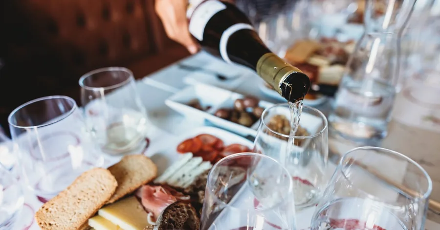 Uma garrafa a servir vinho branco em copos ao lado de uma tábua com queijo e enchido